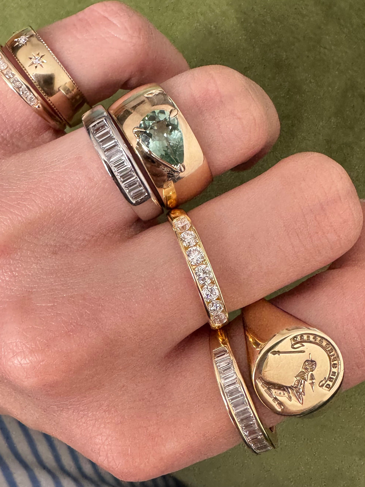 A close-up of a hand wearing multiple rings, including yellow gold signet rings, gold bands, silver rings with baguette diamonds, and a ring with a green emerald-cut stone, set against an olive green background.