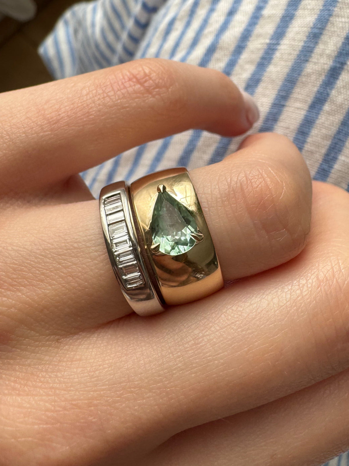 A close-up of a hand wearing two rings: a white gold band with baguette diamonds, and a yellow gold band with a triangular-cut green tourmaline