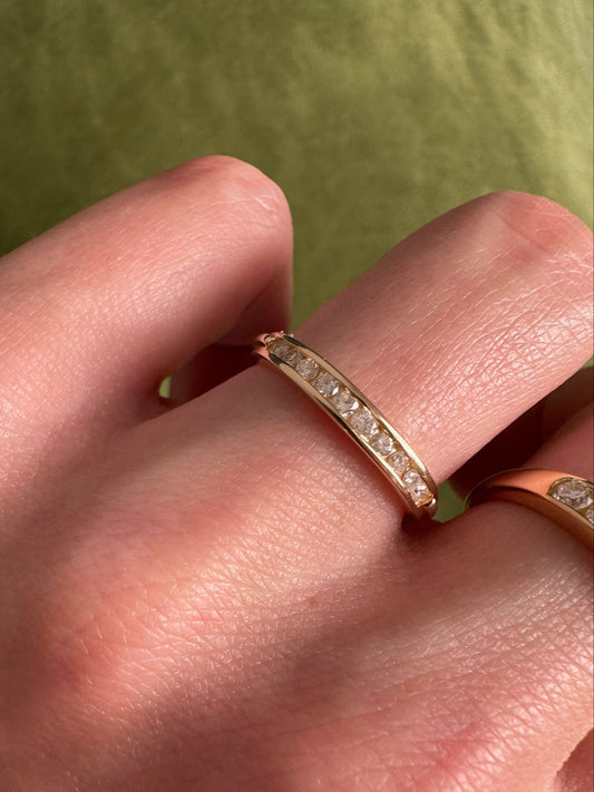 A close-up of a hand wearing a slim gold band ring with a row of small diamonds, with another gold ring partially visible on the same finger